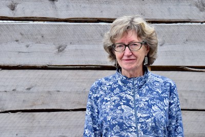 Photo of Nan Heald, an older white woman with dark and graying hair, wearing glasses. She is standing in front of a wooden backdrop, wearing a blue patterned top and smiling. 
