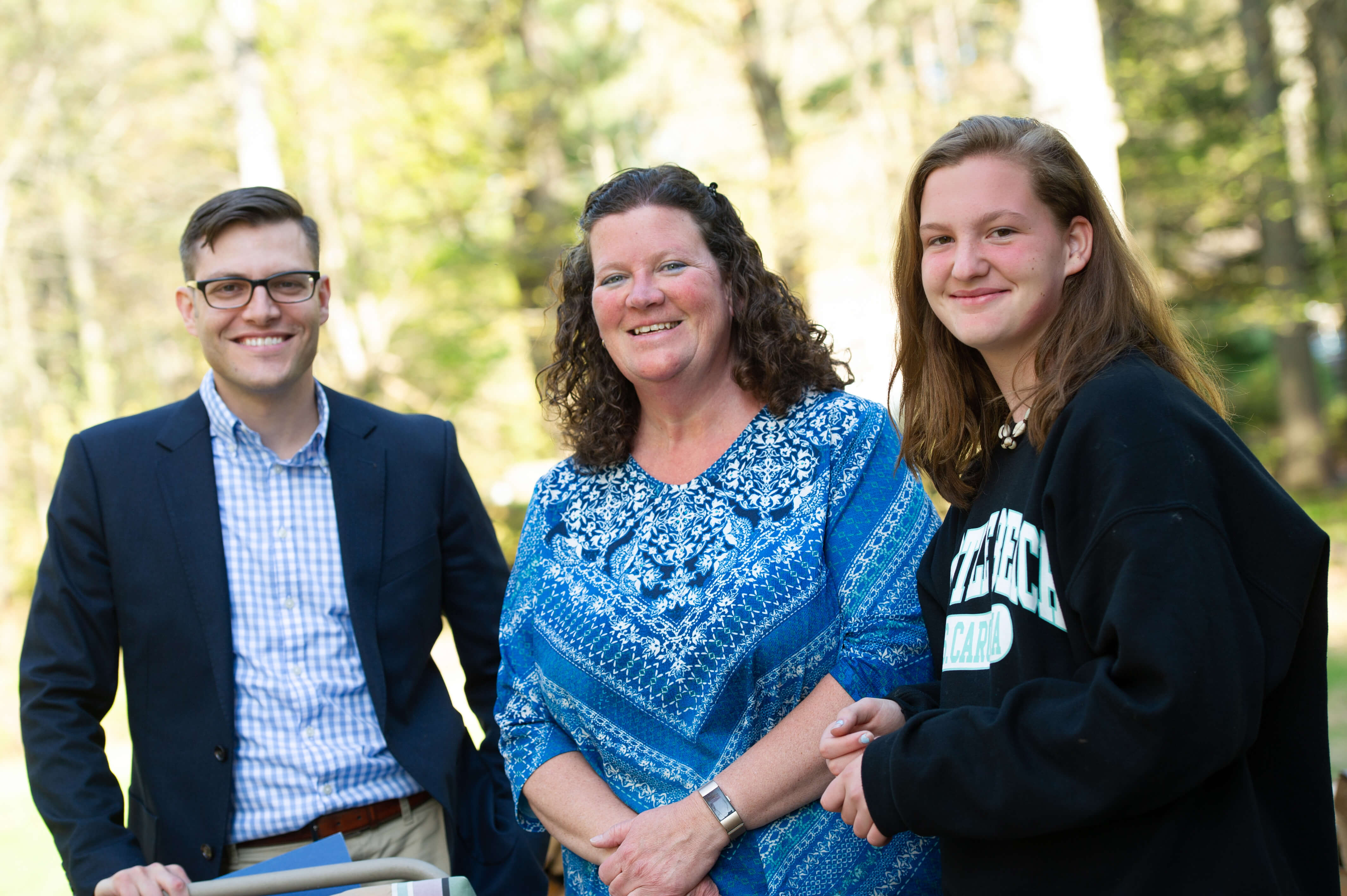 Three white people stand in a line, looking to camera and smiling. On the left, a man with short brown hair and glasses, wearing a button-up shirt and suit coat. In the middle, a woman with shoulder-length brown hair, wearing a patterned blue top. On the right, a teenage girl with shoulder length brown hair, wearing a dark hoodie.