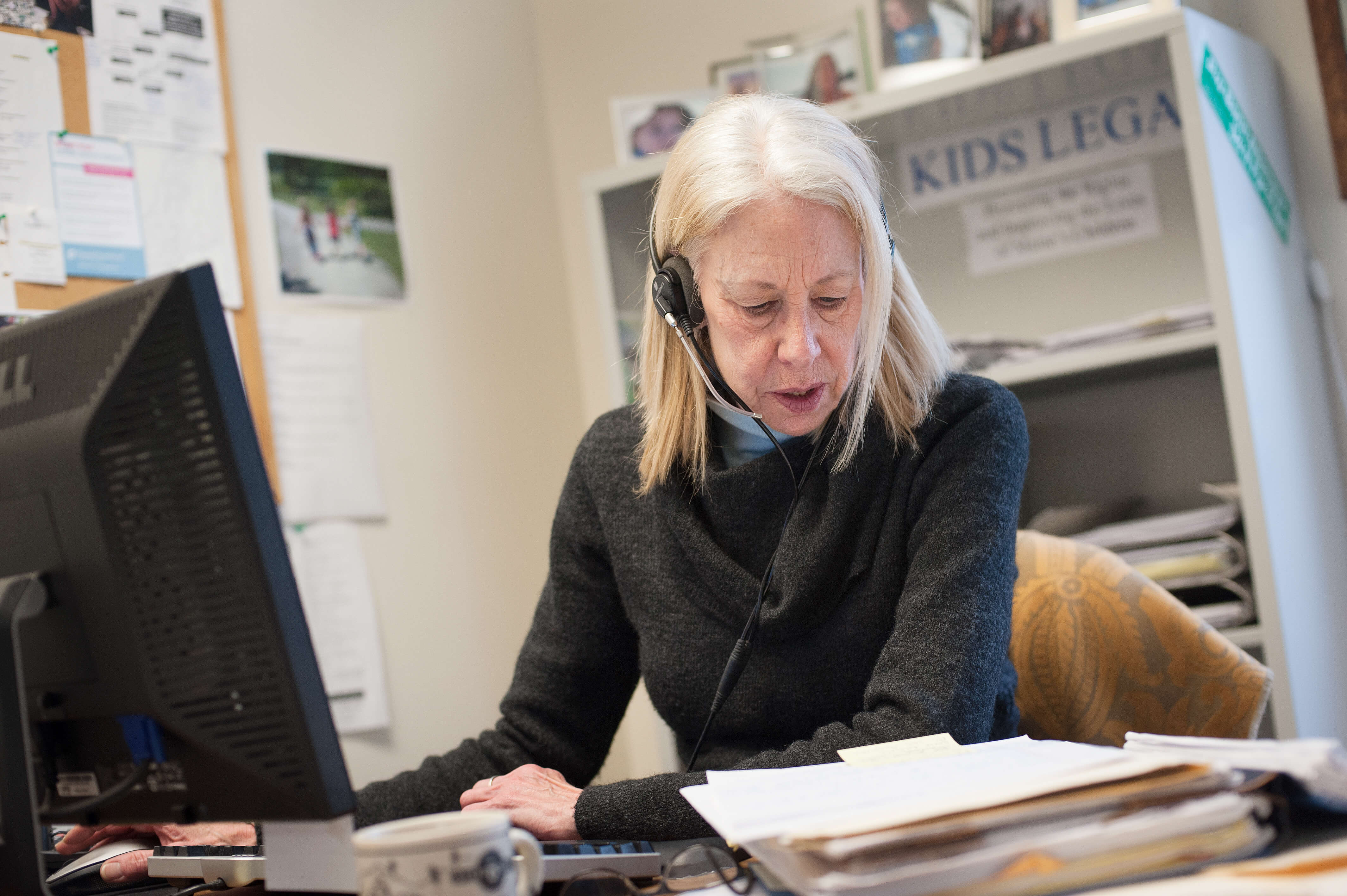 An older white woman with shoulder-length blonde hair sits inan office, in front of a computer. She is wearing a phone headset and appears to be talking with someone while reviewing a stack of documents.