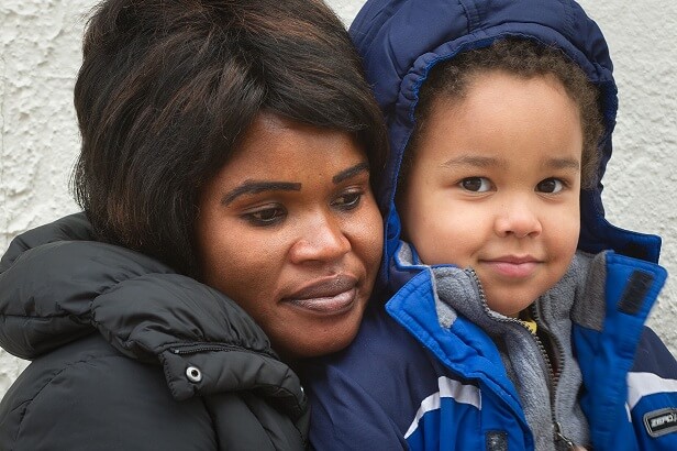 A Black woman with short, straight black hair, wearing a winter coat holds her toddler son. A young Black boy, in his mother's arms, wearing a blue winter coat, smiles a little at the camera. 
