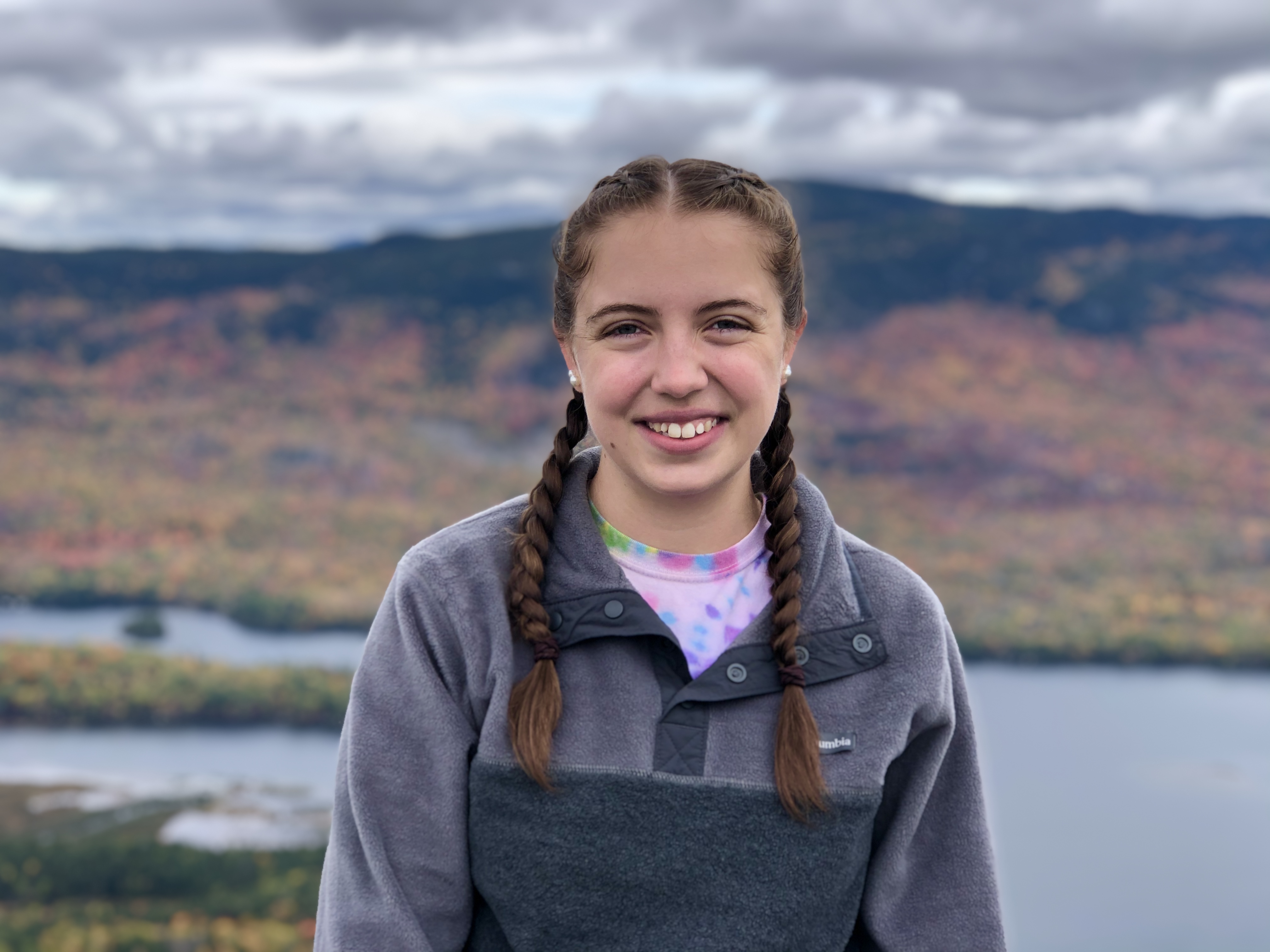 A young white woman stands in the foreground, against a cloudy sky, and in the distance behind her, a lake is framed by a mountain with changing fall foliage. She has long brown hair in braids, she is wearing a gray pullover and smiling at camera. 