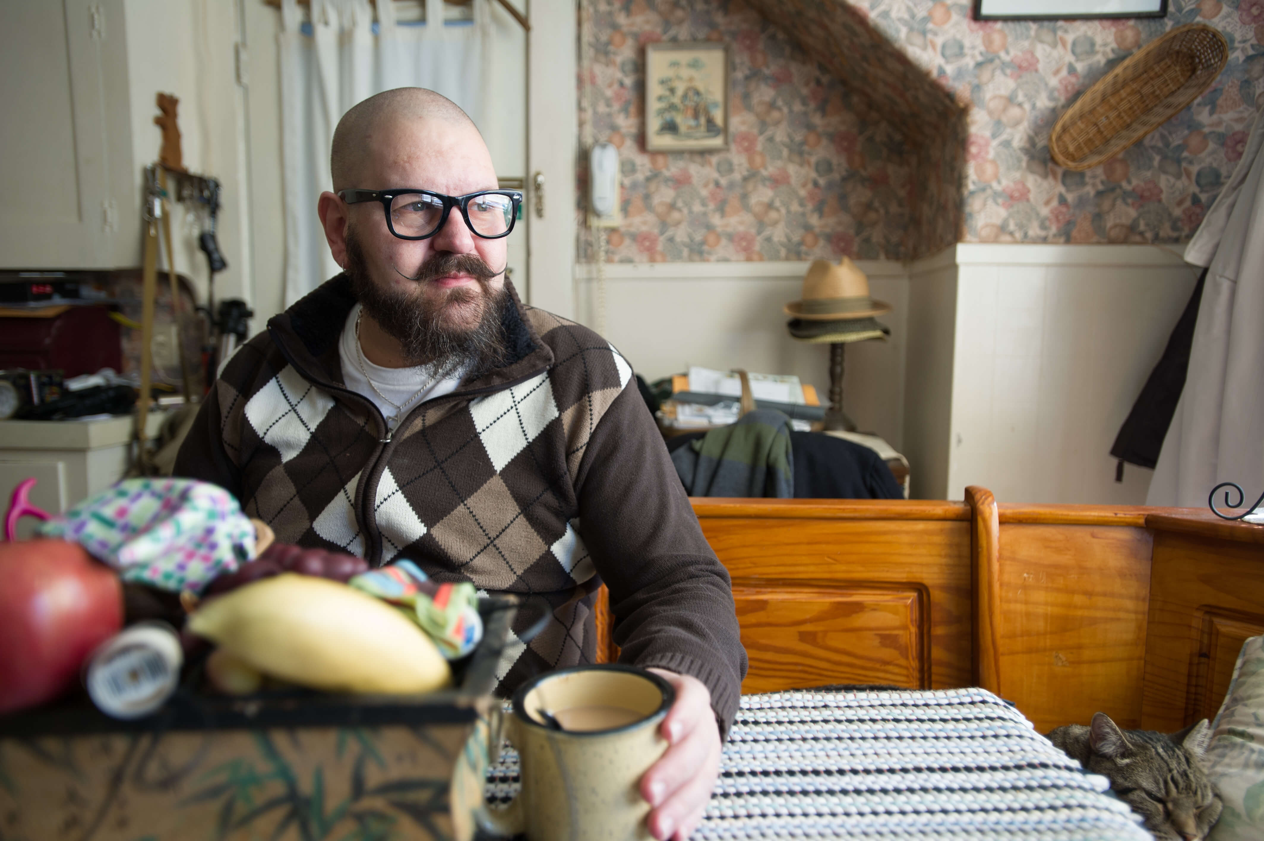 A bald white man with a beard, mustache, and glasses sits at a table in his home, holding a cup of coffee. A tabby cat is curled up next to him. 