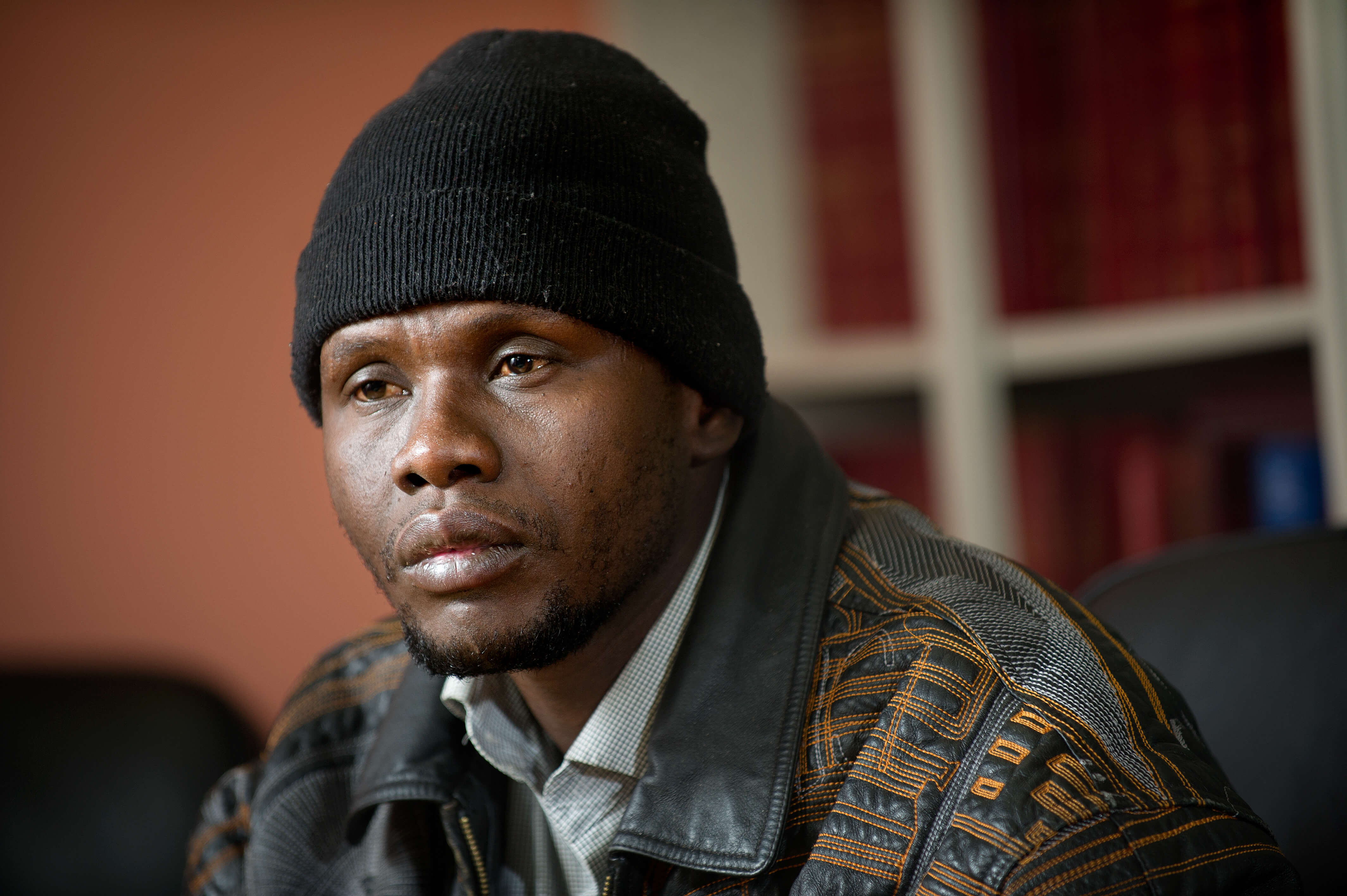 A Black man wearing a leather jacket and black beanie hat sits in an office.