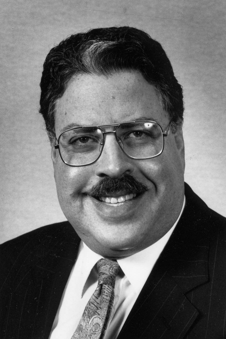 A black-and-white headshot-style photo of a man with light skin, a dark mustache and dark hair with hints of gray. He is wearing glasses, a suit jacket, and tie, and is smiling at the camera. 