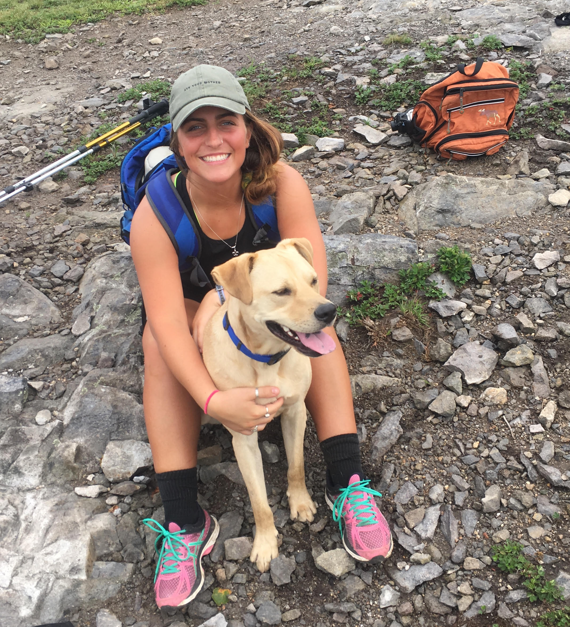 A young white woman in athletic clothing and a baseball cap sits on the ground outdoors. She is smiling at the camera and hugging a yellow dog. 