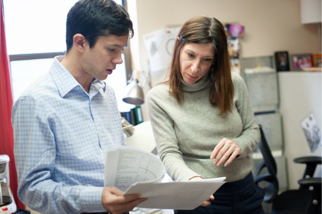 Two white people stand in an office, looking intently at some documents. Left, a man with short brown hair wearing a blue checked button-up shirt. Right, a woman whith long brown hair and a gray sweater. 