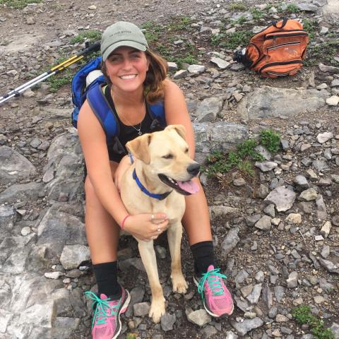 A young white woman sits on the ground hugging her dog. She is wearing a gray baseball hat, a backpack, and hiking clothes, holding on to a yellow lab type dog who is sitting between her feet. She is smiling at camera, and so is the dog.
